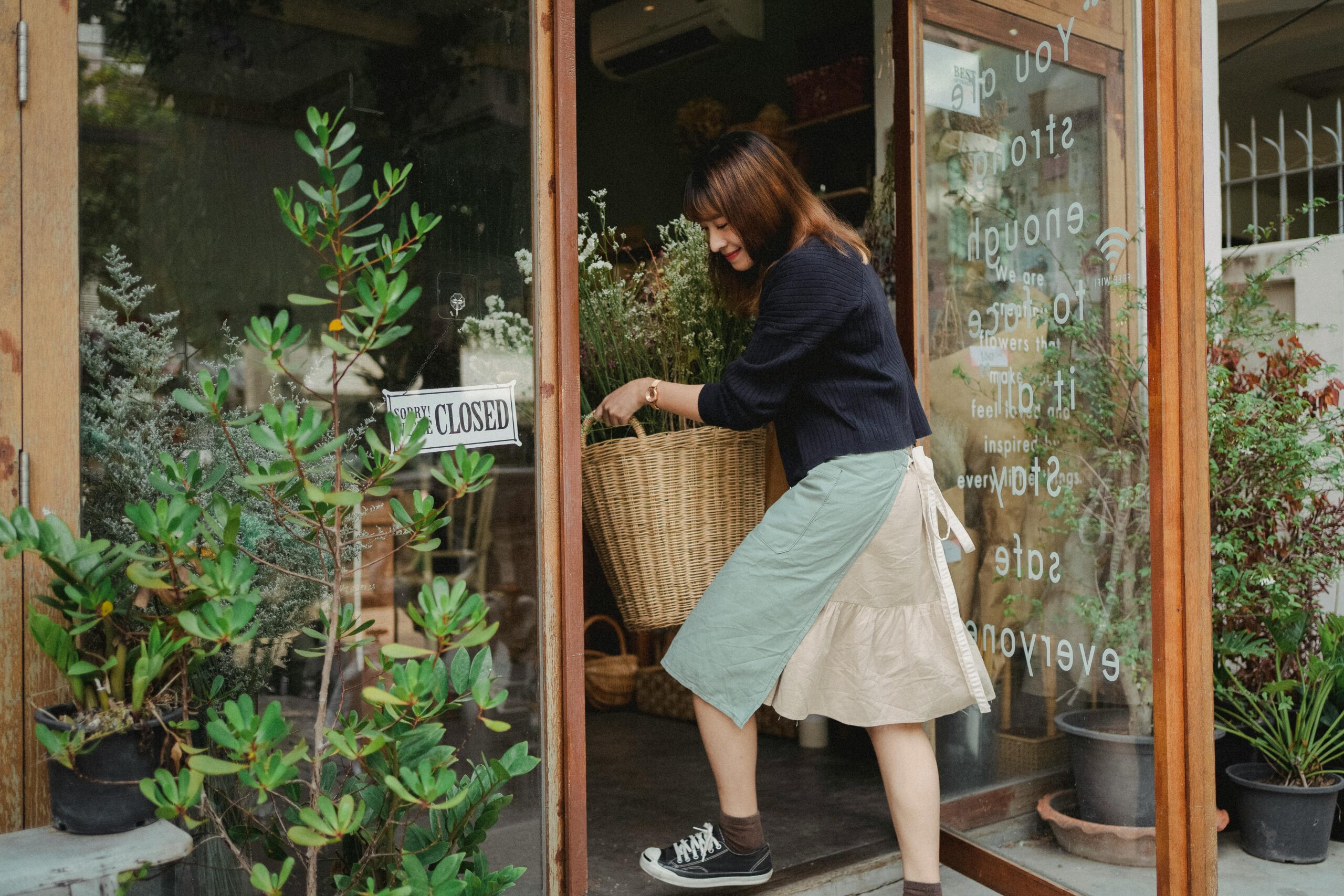 Side view of Asian female florist in casual clothes carrying basket with flowers into shop